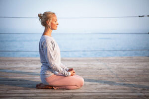 young woman mastering stress by meditating and kneeling with eyes closed on decking in front of sea